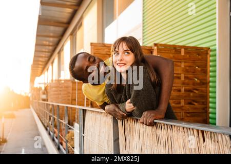 Vue latérale d'un jeune couple multiracial ravie dans une tenue décontractée souriante et serpentant tout en se tenant sur le balcon de la maison et en se penchant sur les balustrades contre les s.