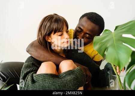 Jeune couple multiethnique attentionné dans des vêtements décontractés et embrassant tout en étant assis ensemble sur un escalier avec plante verte à la maison Banque D'Images