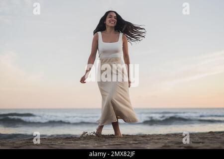 D'en-dessous insouciante jeune hispanique barefooted femme voyageur avec de longs cheveux sombres dans les vêtements d'été souriant tout en marchant sur la plage de sable près de la ageuse f Banque D'Images