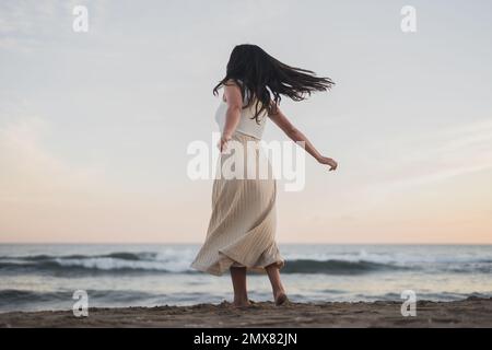 Vue arrière de jeune femme malin et méconnaissable, avec de longs cheveux sombres dans des vêtements d'été dansant sur une plage de sable, près de la poulamière Banque D'Images