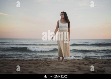 D'en-dessous insouciante jeune hispanique barefooted femme voyageur avec de longs cheveux sombres dans les vêtements d'été debout regardant loin sur la plage de sable près de la ageuse f Banque D'Images