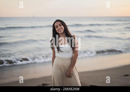 Jeune femme hispanique, insouciante et ébatée, avec de longs cheveux foncés dans des vêtements d'été souriants, debout regardant l'appareil photo sur une plage de sable à proximité Banque D'Images