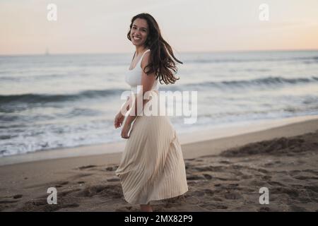 Vue latérale d'une jeune femme hispanique sans soucis, ébouillantée, avec de longs cheveux sombres dans des vêtements d'été souriants tout en regardant l'appareil photo sur le sable Banque D'Images