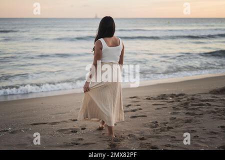 Vue arrière de jeune femme malin et méconnaissable, avec de longs cheveux foncés dans des vêtements d'été, debout sur une plage de sable, près de la mousse Banque D'Images