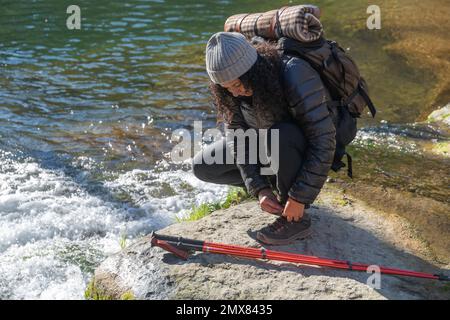 Ensemble complet de femme voyageur dans des vêtements chauds et chapeau avec sac à dos assis sur la rive rocheuse près de l'eau et des bâtons de randonnée tout en nouant des lacets de chaussures Banque D'Images