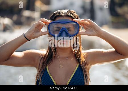 Slim femme plongeur en maillot de bain et lunettes pour plonger sourire et regarder l'appareil photo tout en se relaxant le week-end d'été sur la plage de Cadaques dans le dos éclairé Banque D'Images