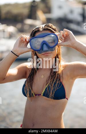 Slim femme plongeur en maillot de bain et lunettes pour plonger sourire et regarder l'appareil photo tout en se relaxant le week-end d'été sur la plage de Cadaques dans le dos éclairé Banque D'Images