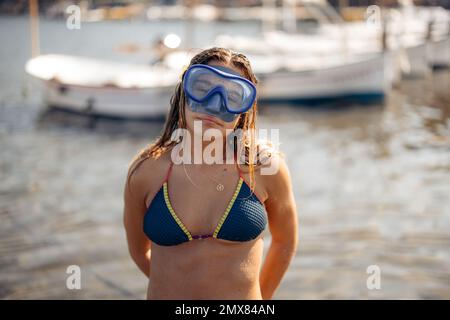 Slim femme plongeur en maillot de bain et lunettes pour plonger sourire et regarder l'appareil photo tout en se relaxant le week-end d'été sur la plage de Cadaques dans le dos éclairé Banque D'Images