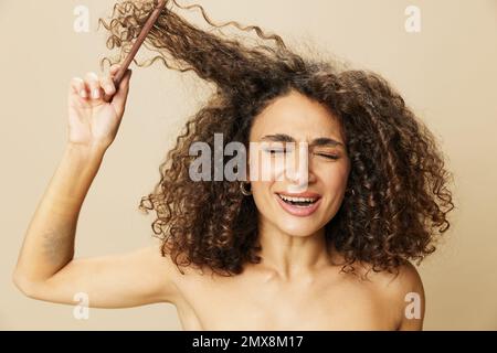 Femme peigne les cheveux bouclés afro avec peigne en bois, produits de coiffage de soins de beauté maison, perte de cheveux après Covid-19, sourire sur fond beige Banque D'Images