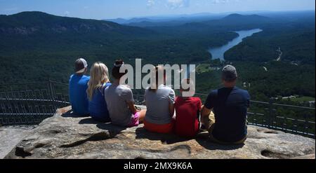 Une famille de six personnes s'assoit au bord du rocher en admirant la vue depuis Chimney Rock, le parc national de Chimney Rock, en Caroline du Nord. Le lac Lure est visible à distance. Banque D'Images