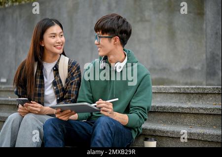 Deux jeunes amis asiatiques heureux d'université assis sur les escaliers de rue, en utilisant une tablette, aiment parler et discuter de leurs projets scolaires. Banque D'Images