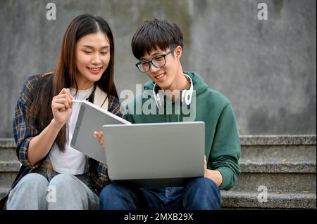 Un jeune étudiant d'université asiatique intelligent utilise un ordinateur portable, parle et travaille sur un projet scolaire avec sa petite amie à l'escalier de rue du bâtiment du campus Banque D'Images