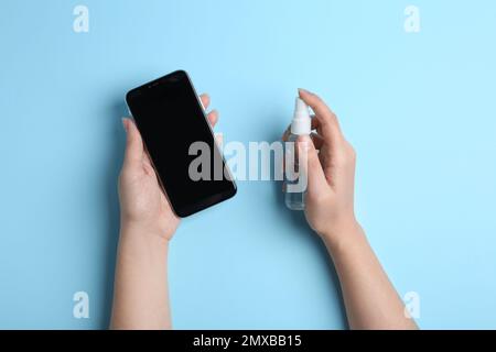 Femme pulvérisant un antiseptique sur un smartphone sur fond bleu clair, vue de dessus Banque D'Images