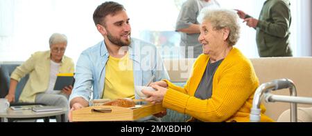 Jeune homme servant dîner pour femme âgée dans un hospice gériatrique, bannière design. Soins aux personnes âgées Banque D'Images