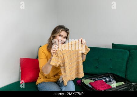 Une femme souriante tient dans les mains un t-shirt rayé assis sur un canapé dans la chambre à la maison. Une femme heureuse replie ses vêtements de loisirs dans ses bagages. Banque D'Images