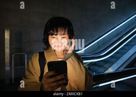 Portrait de la belle fille coréenne dans un casque, se tient près de l'escalier roulant, voyage, se rend à la maison dans les transports publics, tient le smartphone, pose avec mobil Banque D'Images