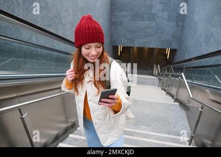 Photo en extérieur d'une jeune femme qui planifie un itinéraire, suit la carte sur l'application pour smartphone, monte les escaliers avec un sac à dos et sourit. Banque D'Images