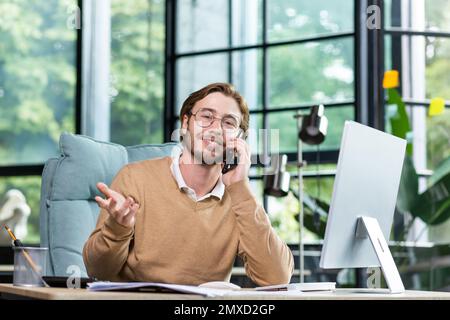 Jeune homme d'affaires beau, indépendant assis dans le bureau à l'ordinateur et parlant au téléphone. Regarde dans l'appareil photo. gestuelle, sourire. Banque D'Images