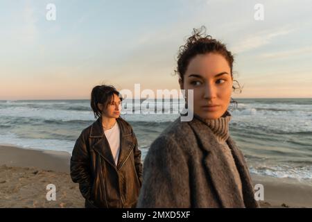 Femme souriante regardant loin près d'un ami flou sur la plage pendant le coucher du soleil à Barcelone Banque D'Images