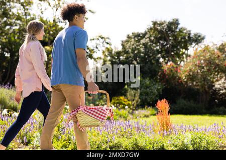 Heureux couple divers tenant les mains marchant dans le jardin ensoleillé avec panier de pique-nique, avec l'espace de copie Banque D'Images