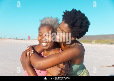 Jeune femme afro-américaine joyeuse embrassant la mère senior et appréciant à la plage contre le ciel bleu Banque D'Images