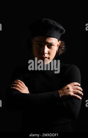 homme afro-américain élégant en beret en col roulé debout avec des bras croisés et regardant la caméra isolée sur noir, image de stock Banque D'Images