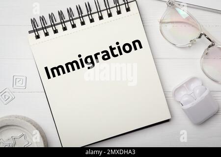 Carnet avec TEXTE IMMIGRATION, lunettes et écouteurs sur table en bois blanc, plat Banque D'Images
