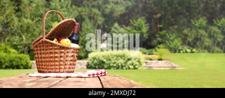 Panier pique-nique avec fruits, bouteille de vin et couverture à carreaux sur table en bois dans le jardin, espace pour le texte. Bannière Banque D'Images