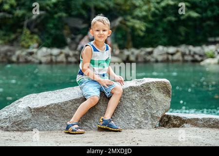 L'enfant excité s'assoit sur le rocher près d'un étang turquoise éclairé par la lumière du soleil. Un garçon d'âge préscolaire souriant et qui aime explorer des endroits magiques dans la station balnéaire Banque D'Images