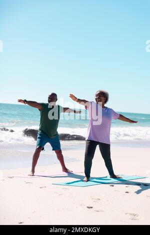 Couple senior afro-américain pratiquant le guerrier 2 pose sur des tapis d'exercice à la plage sous ciel clair Banque D'Images