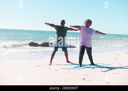 Couple afro-américain senior pratiquant le guerrier 2 pose sur des tapis de yoga à la plage sous ciel clair Banque D'Images