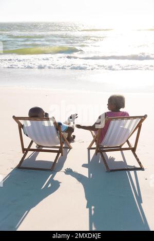 Vue arrière du couple senior afro-américain assis sur des transats à la plage en face de la mer sous le ciel Banque D'Images