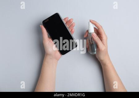 Femme pulvérisant un antiseptique sur un smartphone sur fond gris clair, vue de dessus Banque D'Images