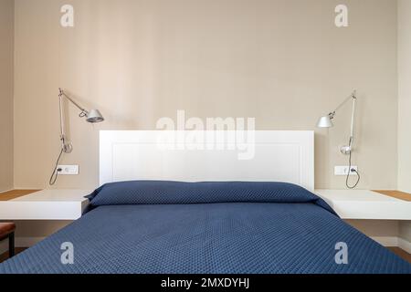 Lit double avec tête de lit blanche recouverte d'un couvre-lit bleu. De chaque côté se trouvent des tables de chevet avec des dessus blancs et des appliques murales avec abat-jour en métal pour Banque D'Images