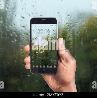 Homme afro-américain utilisant l'application de prévisions météorologiques sur smartphone près de la fenêtre à l'intérieur, gros plan Banque D'Images