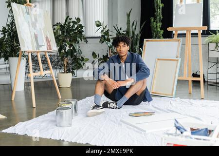 Artiste afro-américain regardant la caméra tout en étant assis près de peintures et de toile sur le sol dans l'atelier, image de stock Banque D'Images