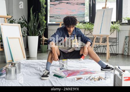 Jeune artiste afro-américain tenant la peinture et le pinceau près de la toile sur le plancher dans l'atelier, image de stock Banque D'Images