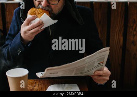 Un barbu d'âge moyen mange un savoureux hamburger et lit le journal d'aujourd'hui dans une rue de la ville en hiver. Concept de cuisine de rue. Banque D'Images