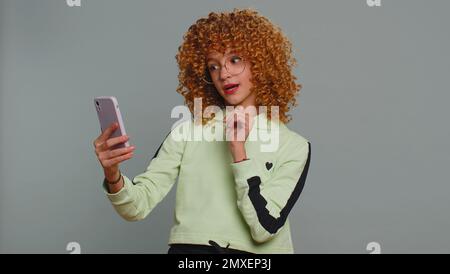 Jeune enfant fille enfant blogueur prenant selfie sur smartphone tenant dans la main, communiquant appel vidéo en ligne avec des abonnés. Adolescents avec cheveux bouclés isolés sur fond gris studio Banque D'Images