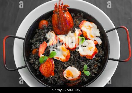 Paella solo avec tranches de homard, morceaux de tomate, sauce à l'encre de calmar, herbes, safran. Plat chaud sur une plaque blanche. Repas sur une table noire. Placage alimentaire moderne. Banque D'Images