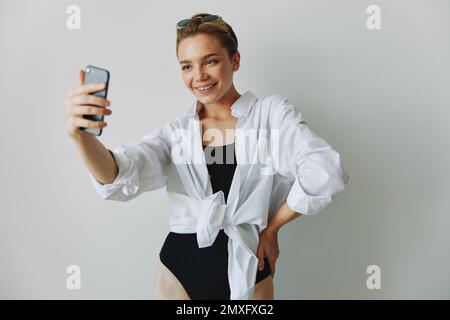 Une jeune fille blogger prend des photos et des pousses. elle-même sur la vidéo sur son téléphone pour le contenu de médias sociaux, blogueur vidéo, se selfie contre un mur blanc avec Banque D'Images