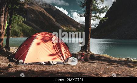 Une tente orange bordée de pierres et d'objets touristiques se dresse dans les arbres sur la rive du lac alpin Shavlinskoye en Altaï après une pluie Banque D'Images