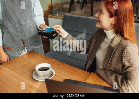 Femme payant par carte de crédit avec NFC dans un café. Femme aux cheveux de gingembre assise à la table payant sa facture. Banque D'Images