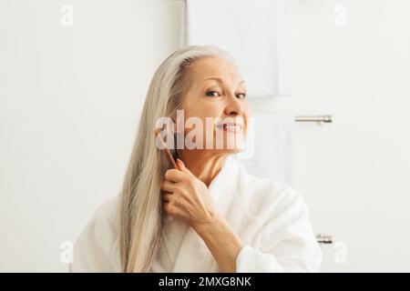 Femme adulte senior peignant ses cheveux avec un peigne en bois. Femme souriante dans un peignoir prenant soin de ses longs cheveux gris. Banque D'Images