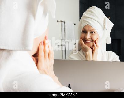 Une femme gaie admire son reflet dans le miroir de la salle de bains. Une femme mûre se touchant le visage avec les mains après le spa à la maison. Banque D'Images