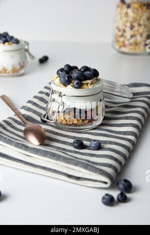 Petit déjeuner flocons d'avoine avec des fruits dans un pot en verre à base de yaourt grec et granola garni de myrtilles fraîches sur un tissu rayé avec cuillère à miel, santé Banque D'Images