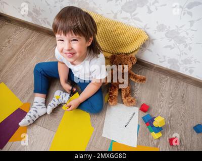 Le jeune garçon apprend à couper du papier de couleur avec des ciseaux. Les enfants sont assis au sol dans la chambre des enfants avec des blocs de jouets et un ours en peluche. Cours d'éducation pour les enfants Banque D'Images
