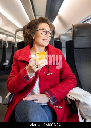 Femme souriante dans un duffle-coat rouge envoyant un SMS sur un smartphone dans un train de banlieue. Voyager en véhicule terrestre. Banque D'Images