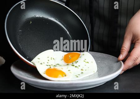 Femme mettant savoureux oeufs cuits sur l'assiette de la poêle à frire à table noire, gros plan Banque D'Images