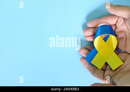 Mains humaines tenant le ruban bicolore bleu et jaune sur fond bleu. Journée mondiale contre le syndrome de Down, sensibilisation, aide et concept de soutien. Banque D'Images